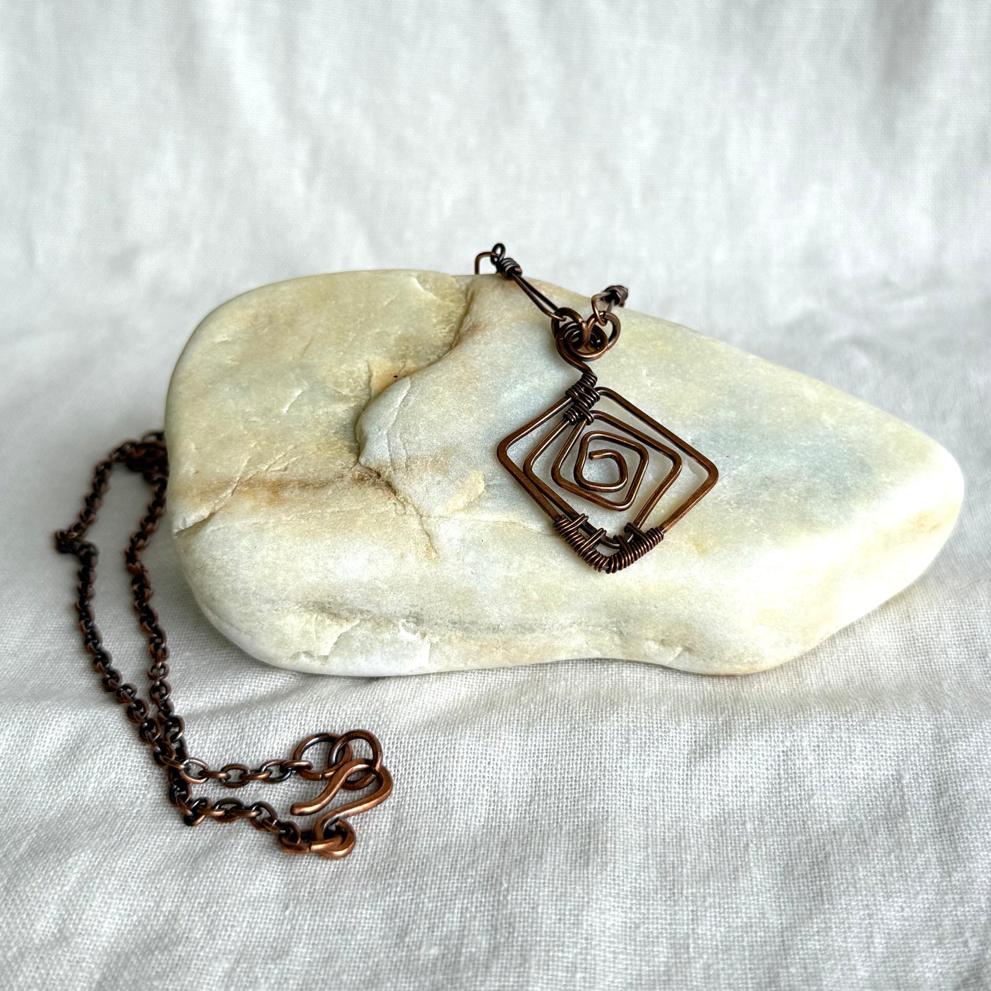 Copper necklace with a geometric pendant on a stone and white fabric background