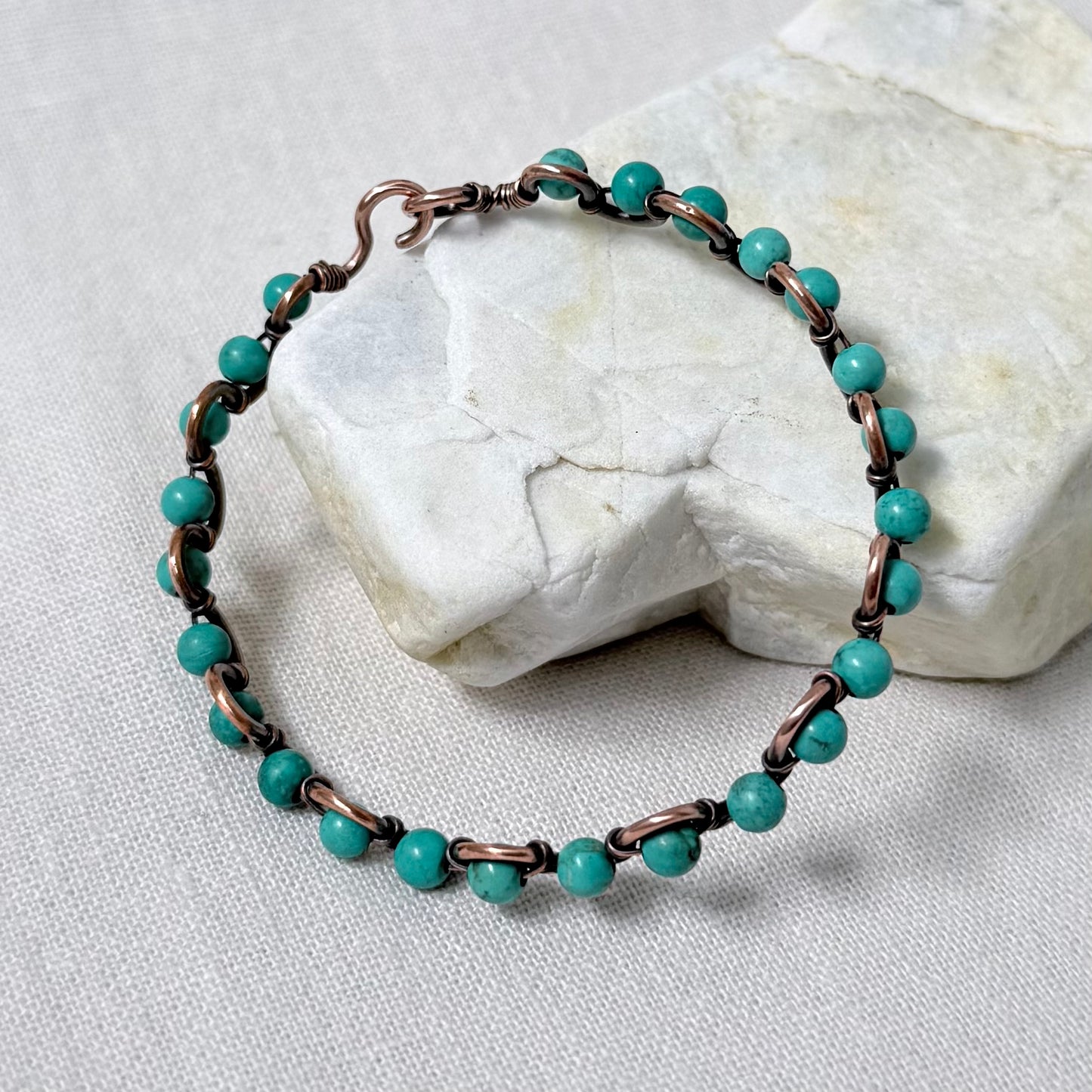 A delicate beaded copper bracelet with sinking turquoise beads, displayed on a light-coloured stone against a neutral fabric background.