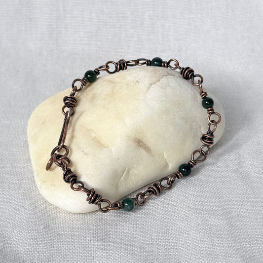 A handmade copper wire bracelet with dark green Moss Agate beads, displayed on a smooth, rounded beige stone against a textured light grey fabric background.