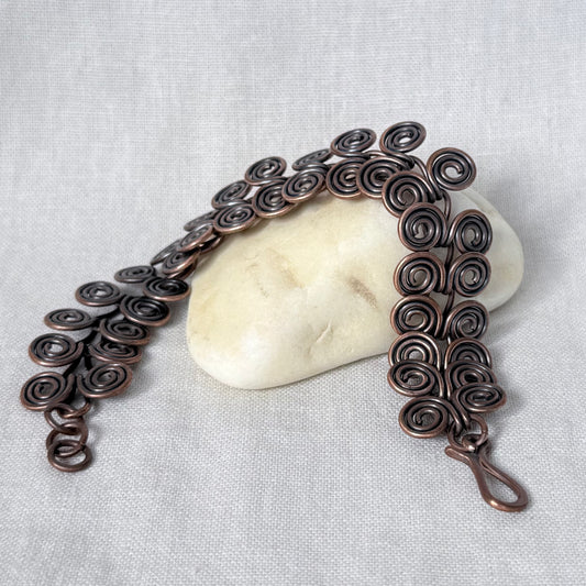 A handmade copper wire chain bracelet with intricate spiral curls, draped over a smooth white stone on a textured light gray fabric background.
