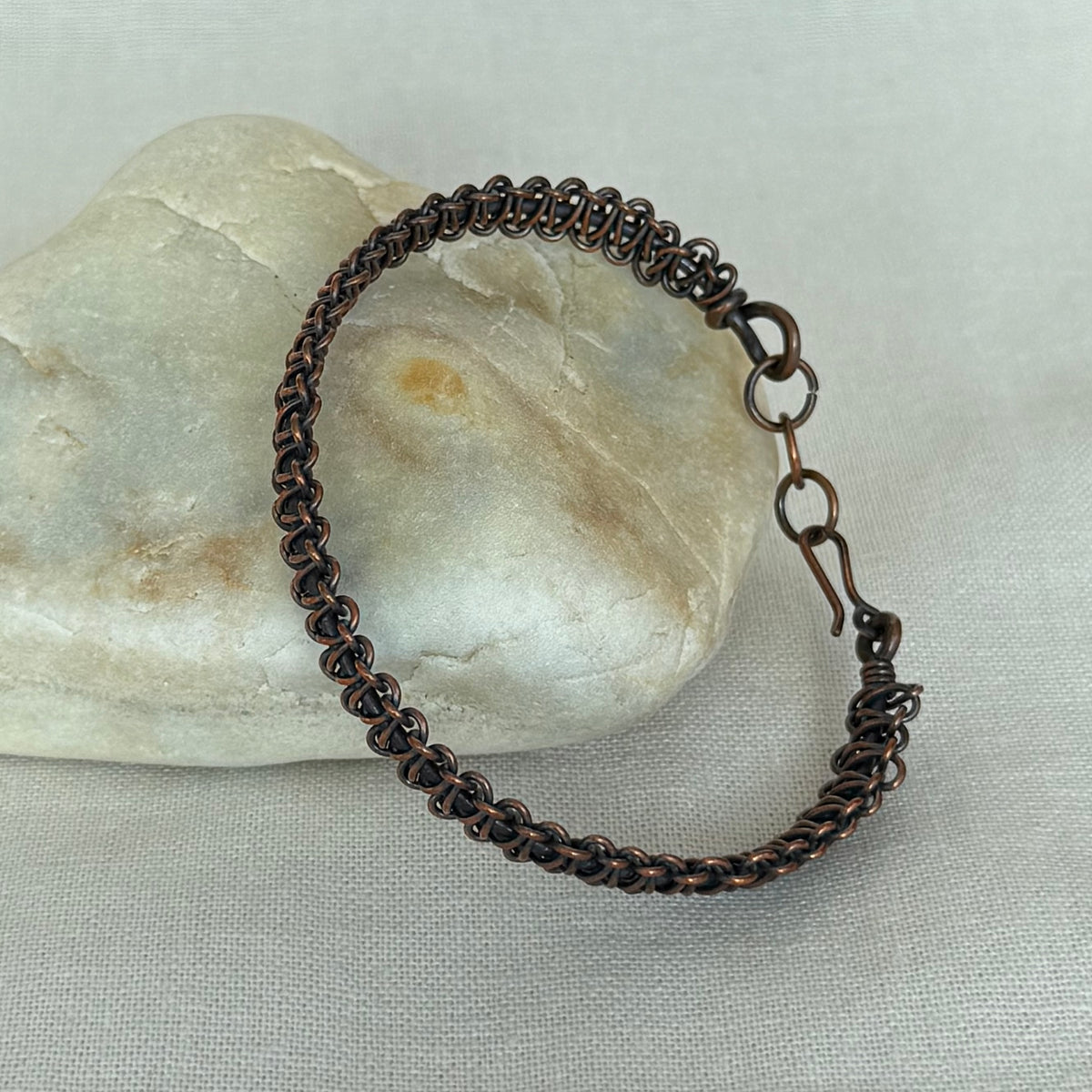 A copper woven bracelet with a coiled spring design and a spring-ring clasp, resting on a smooth white stone over a light grey fabric background.