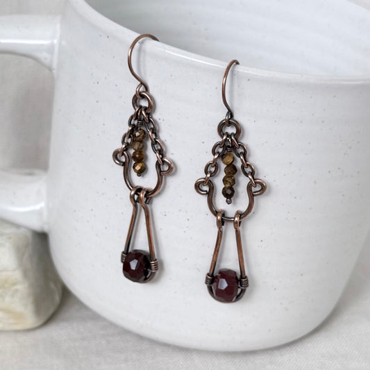A pair of copper drop earrings with small brown beads and a faceted dark red gemstone at the bottom, hanging from the rim of a white speckled ceramic mug.