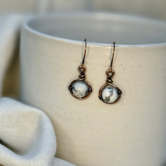 Pair of Copper Earrings with a round howlite Bead hanging from a cream coloured mug on linen background