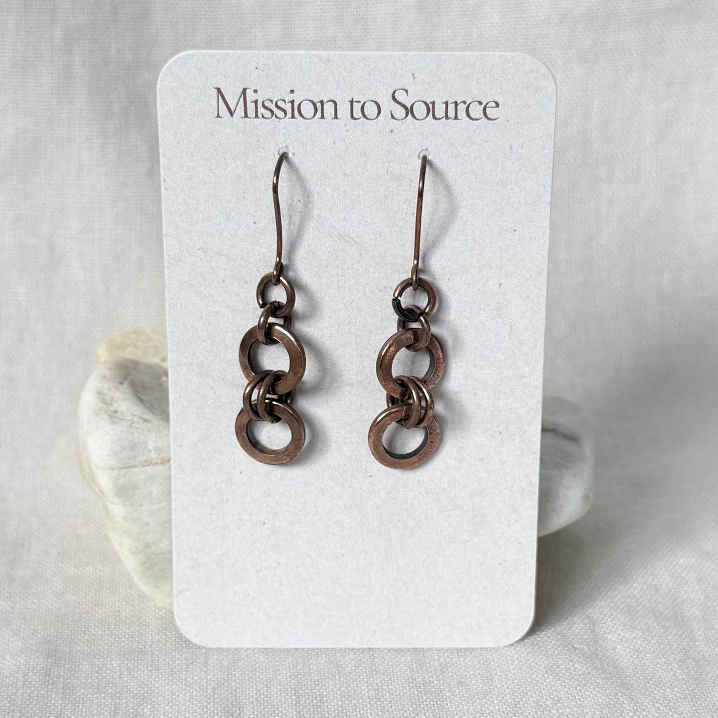 A pair of dangling copper earrings with interlocking circular links, displayed on a white card labeled 'Mission to Source' against a soft neutral fabric background.