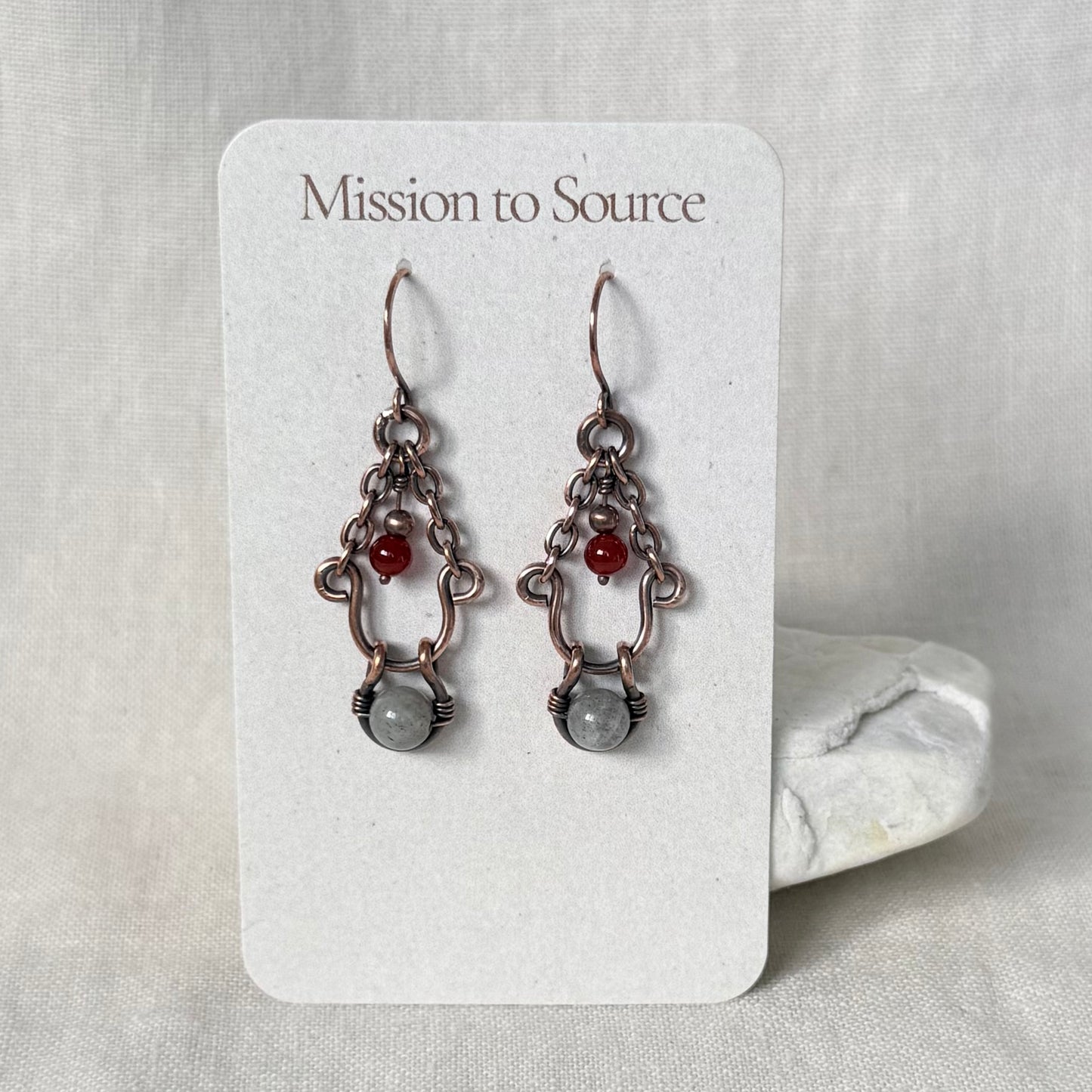 A pair of handmade dangle earrings displayed on a white card labeled 'Mission to Source.' The earrings have copper metalwork with small red beads and a larger pale grey stone at the bottom.