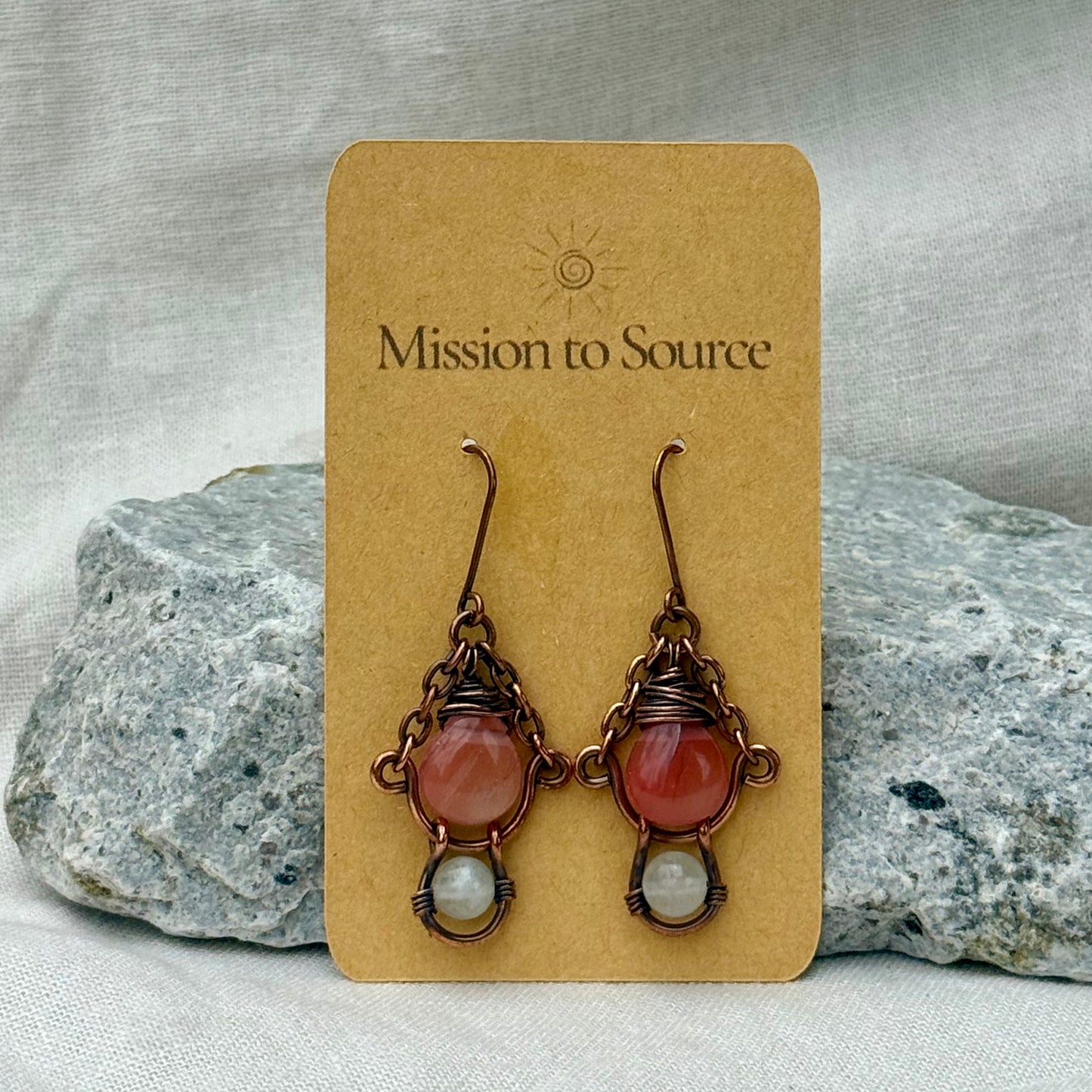 Pair of copper earrings with pink and white beads on a 'Mission to Source' card with rocks in the background