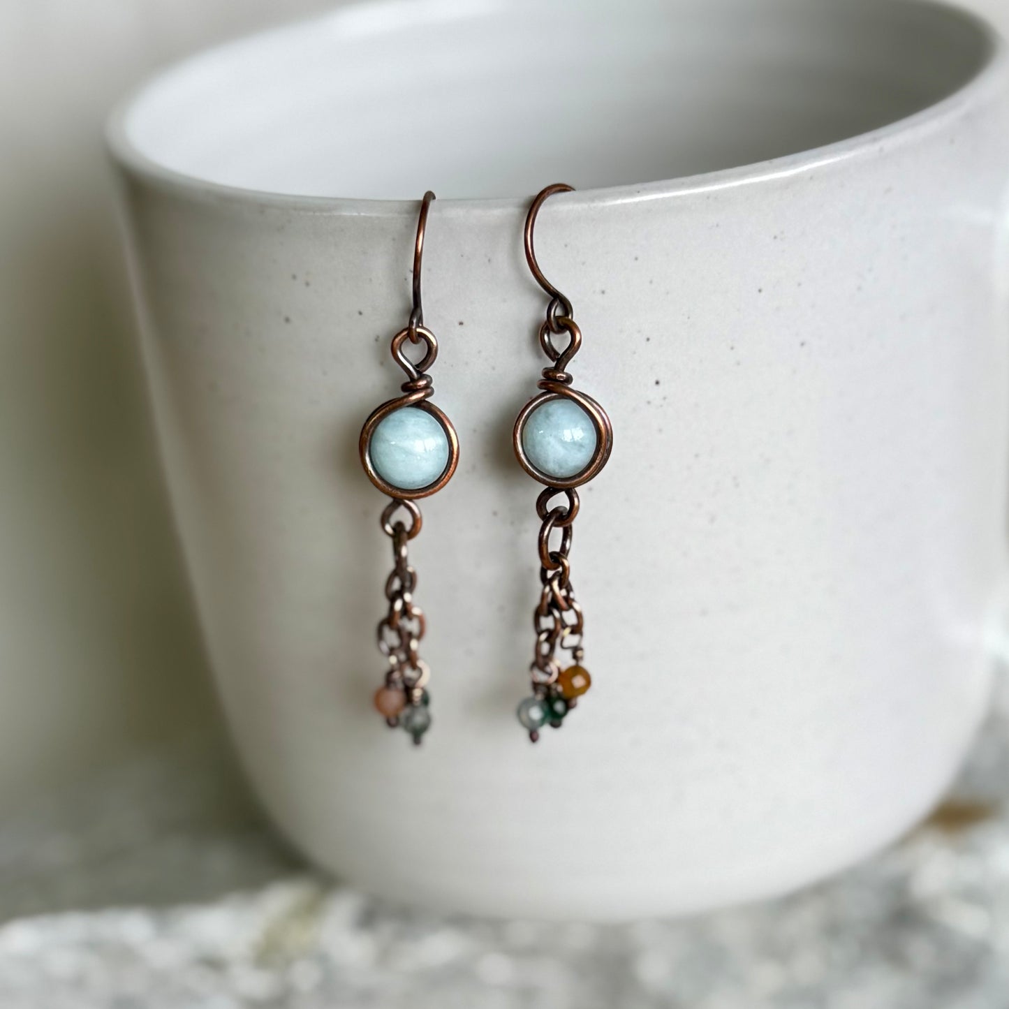 Pair of copper earrings with turquoise stones and chain details on a white ceramic cup.