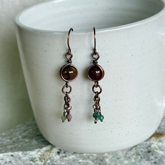 Pair of copper earrings with brown and green gemstones hanging in front of a white ceramic cup.