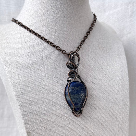 A handmade copper wire-wrapped pendant necklace featuring a blue lapis lazuli stone with gold veining, displayed on a white fabric mannequin bust.