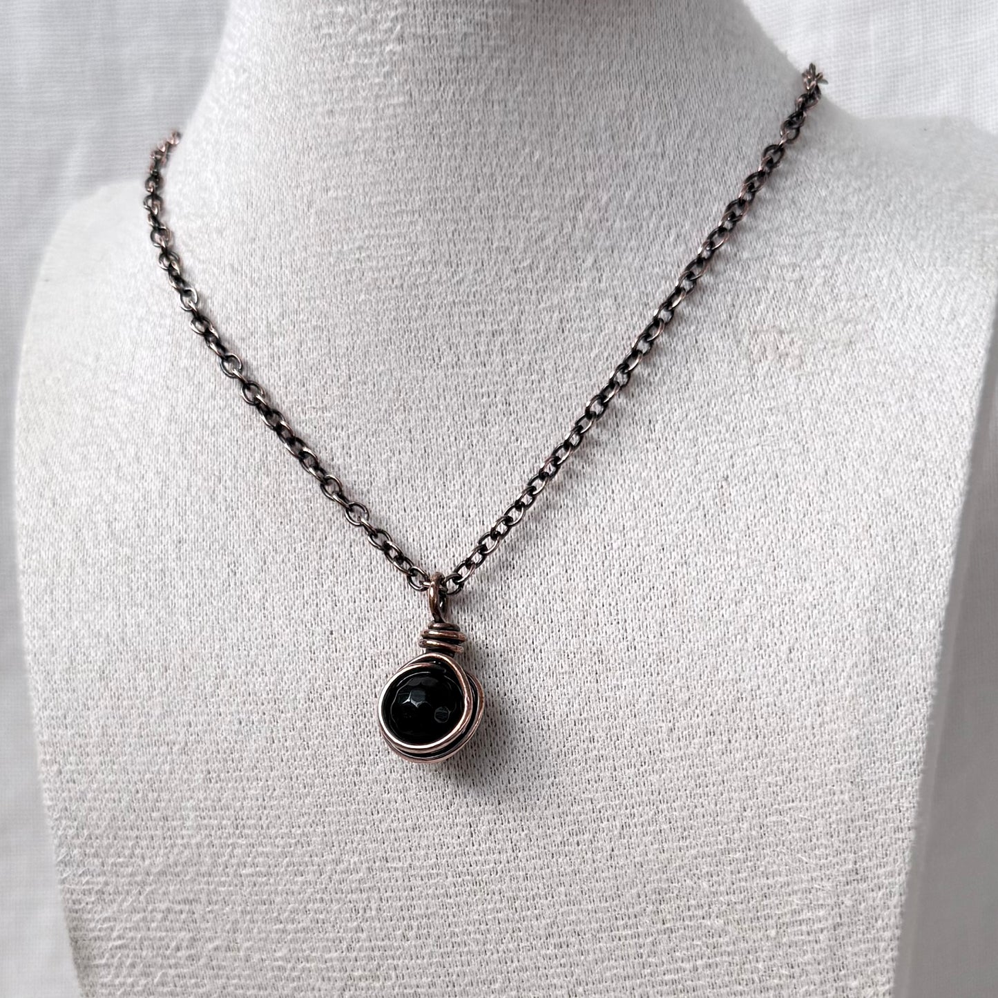 A necklace with a dark round Moss Agate stone in a copper wrap, displayed on a white fabric jewellery bust.