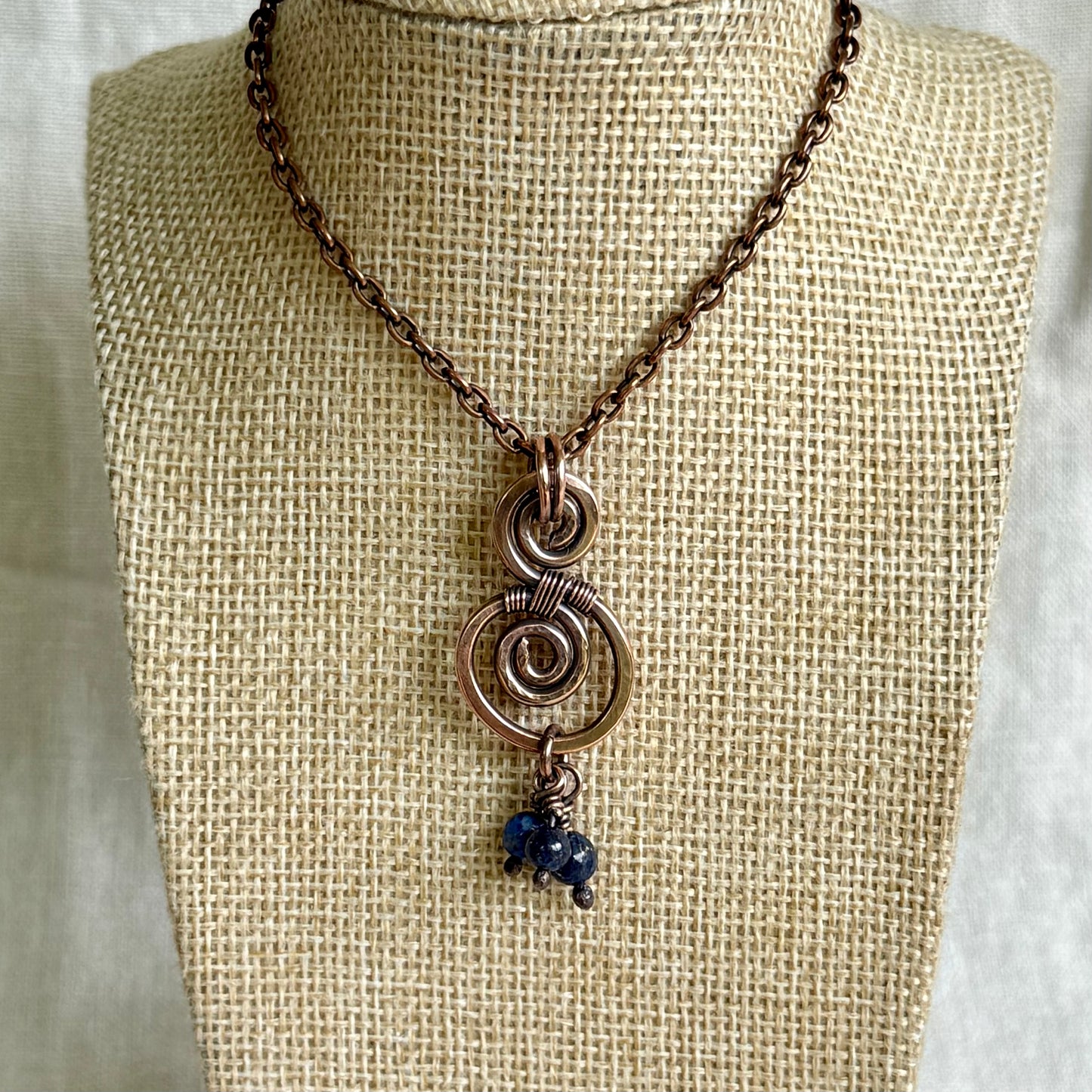 Necklace with a copper spiral pendant and blue beads on a beige mannequin bust.