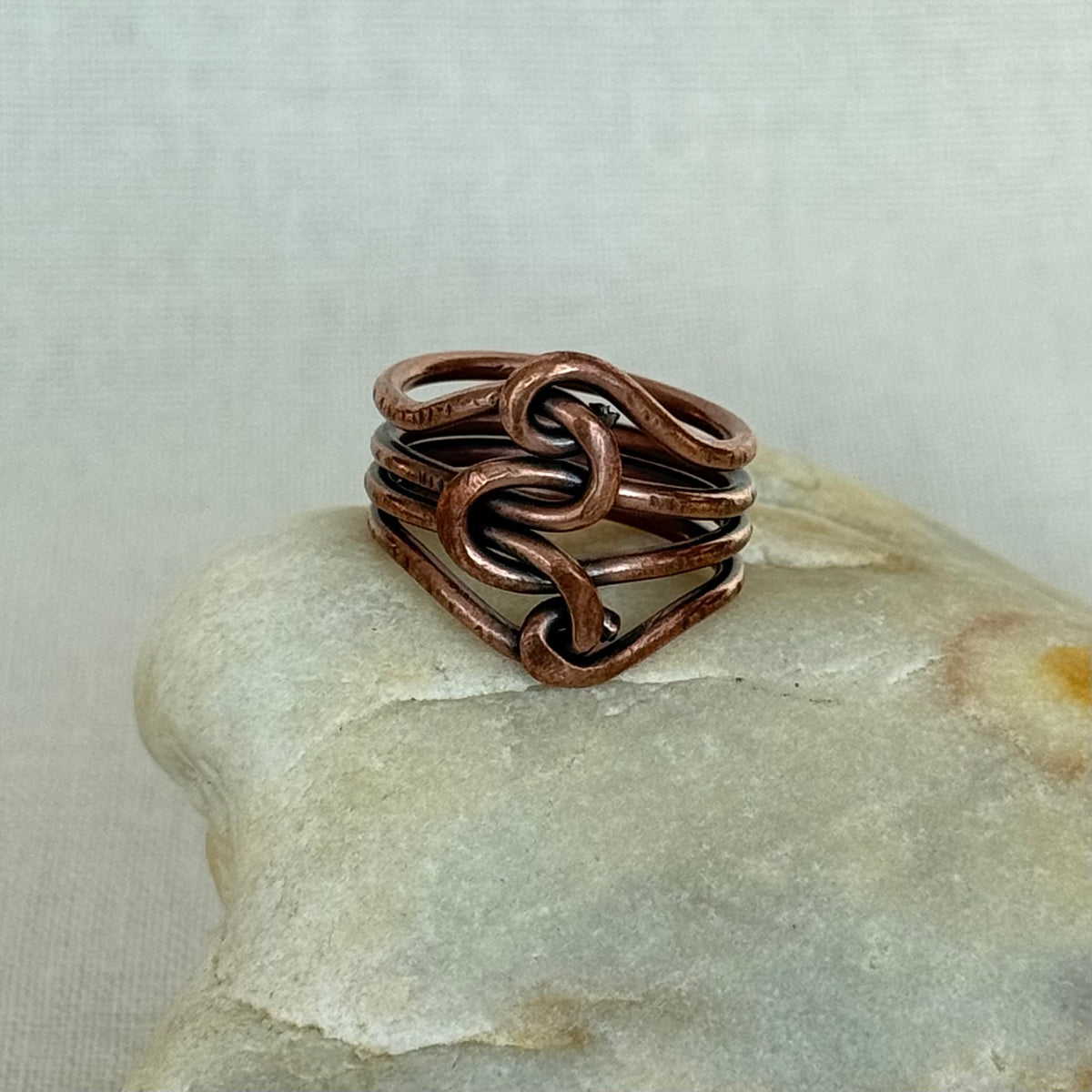 A handmade copper wire ring with a knotted, interwoven design, displayed on a smooth light-colored stone against a neutral textured background.