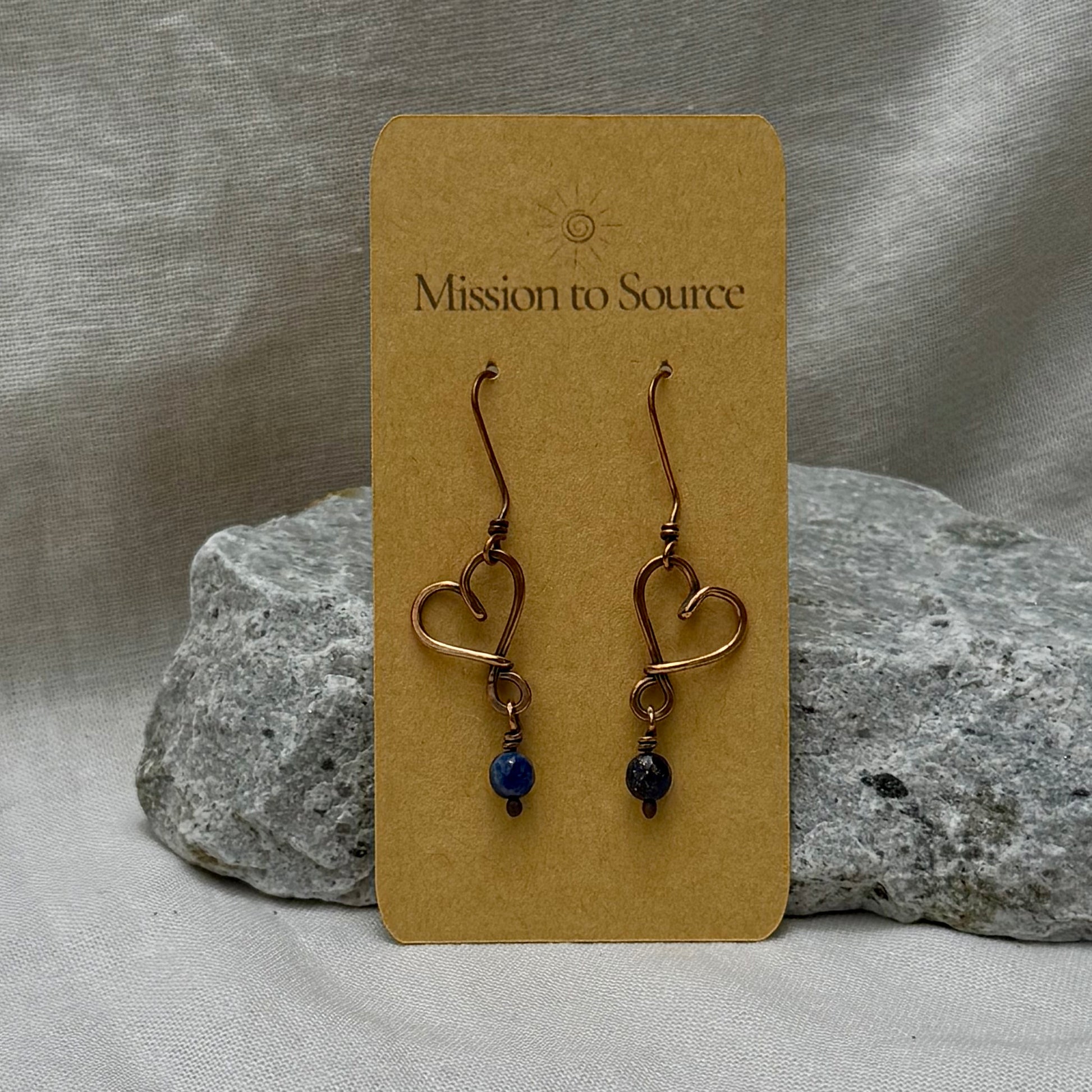 Heart-shaped copper wire earrings with bead on a 'Mission to Source' card with a stone  and linen background