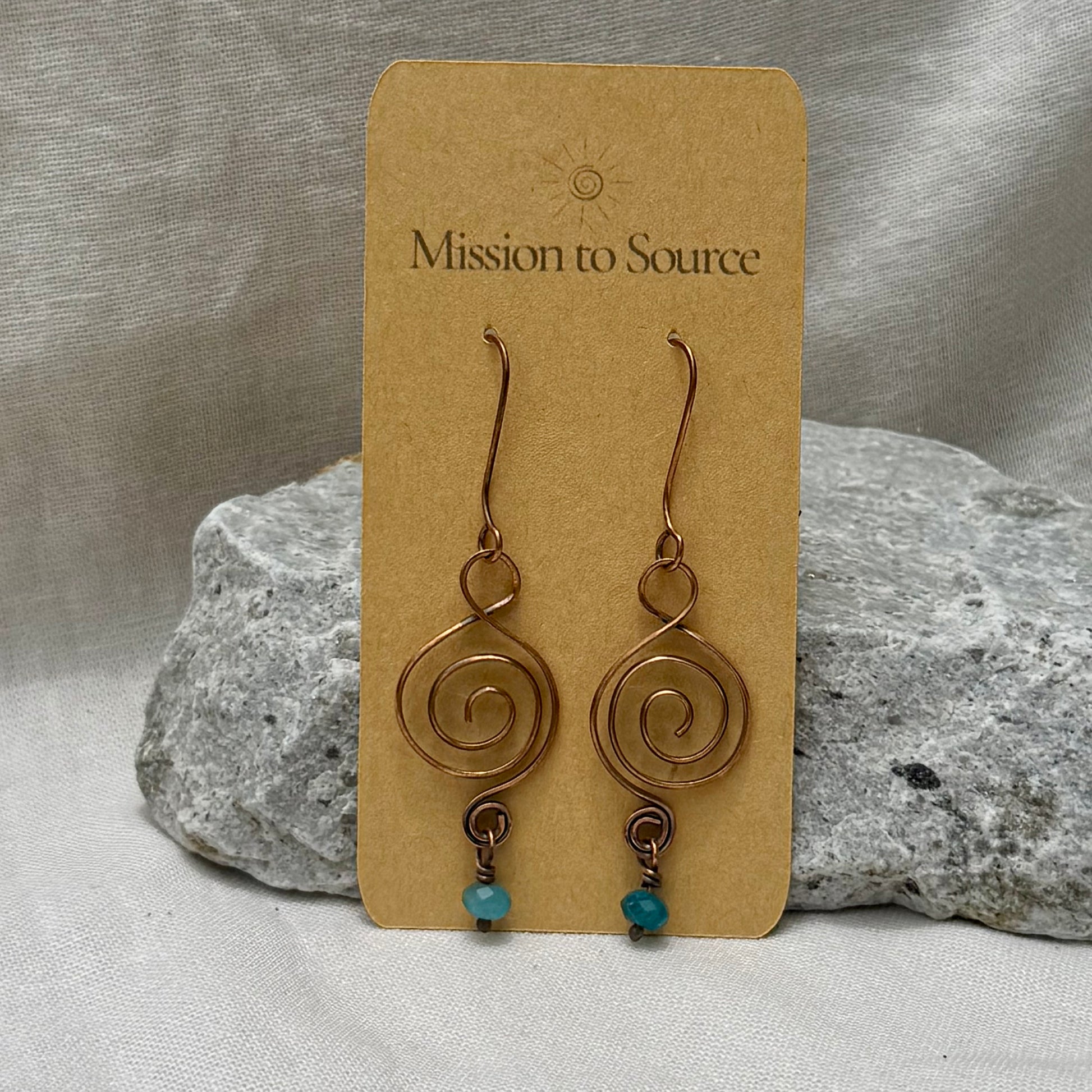 Pair of copper spiral earrings with turquoise beads on a 'Mission to Source' card, placed on a textured surface.