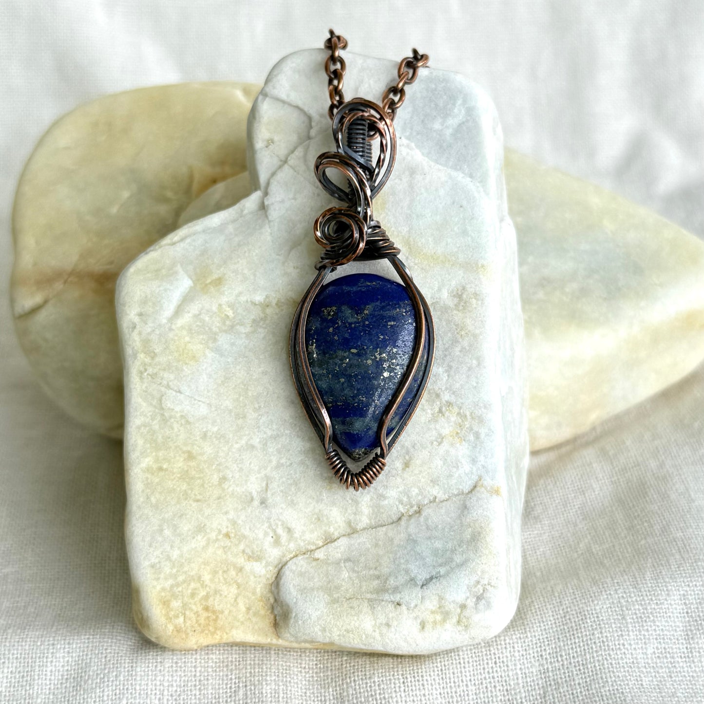 Copper pendant with blue stone and a black cord on a piece of stone on linen background