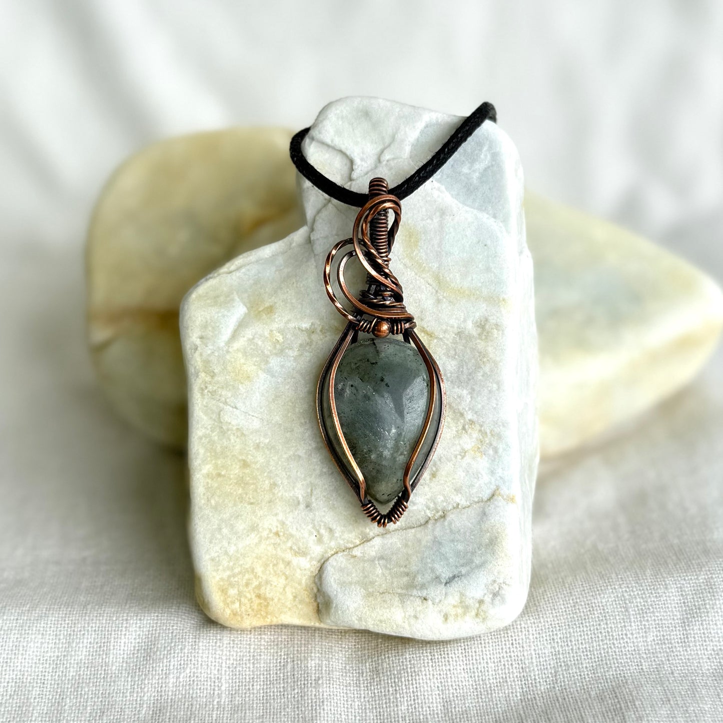 Copper pendant with labradorite stone and a black cord on a piece of stone on linen background
