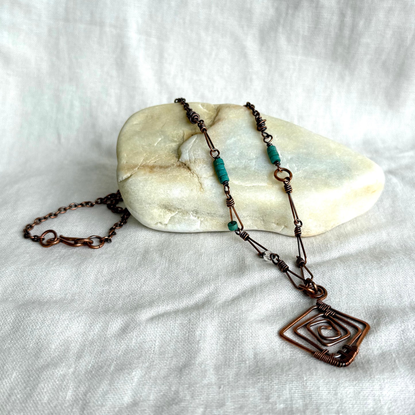 Copper necklace with a geometric pendant on a stone and white fabric background