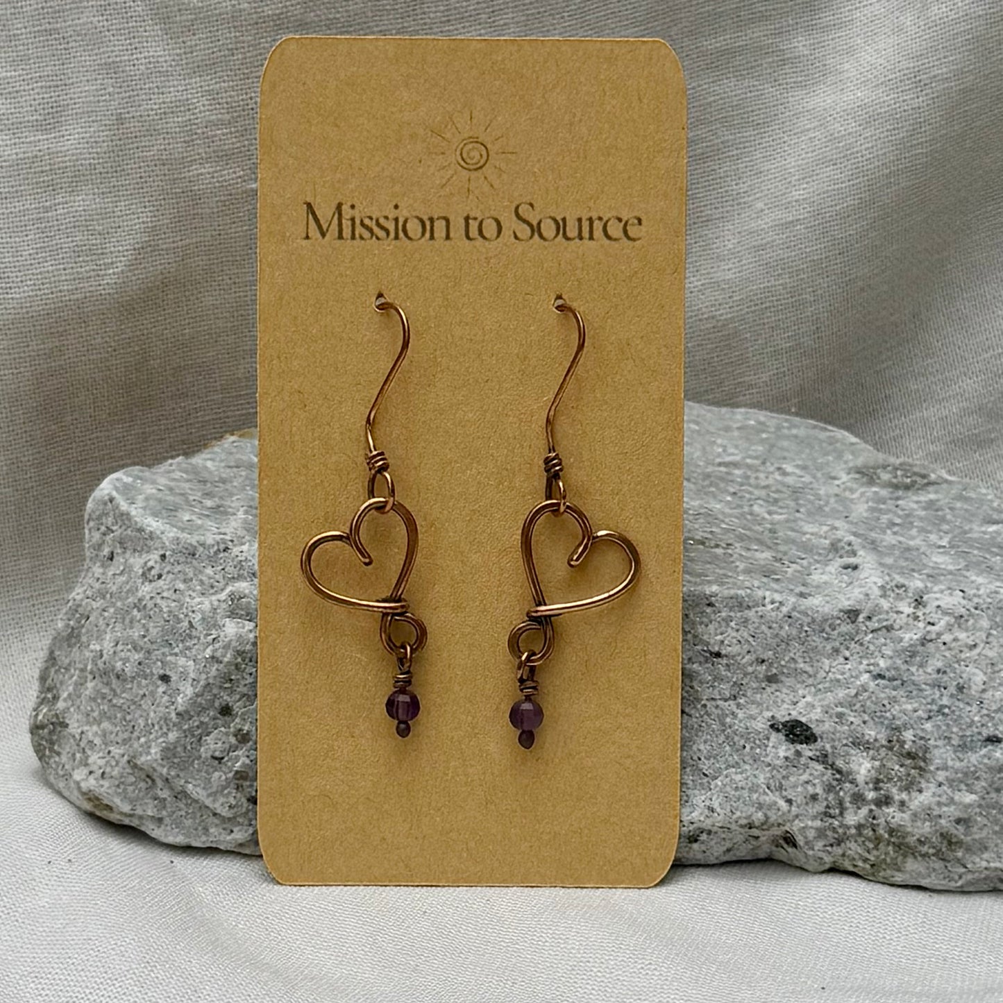 Heart-shaped copper wire earrings with bead on a 'Mission to Source' card with a stone  and linen background
