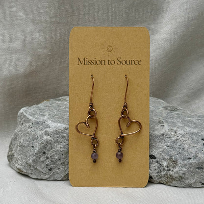 Heart-shaped copper wire earrings with bead on a 'Mission to Source' card with a stone  and linen background