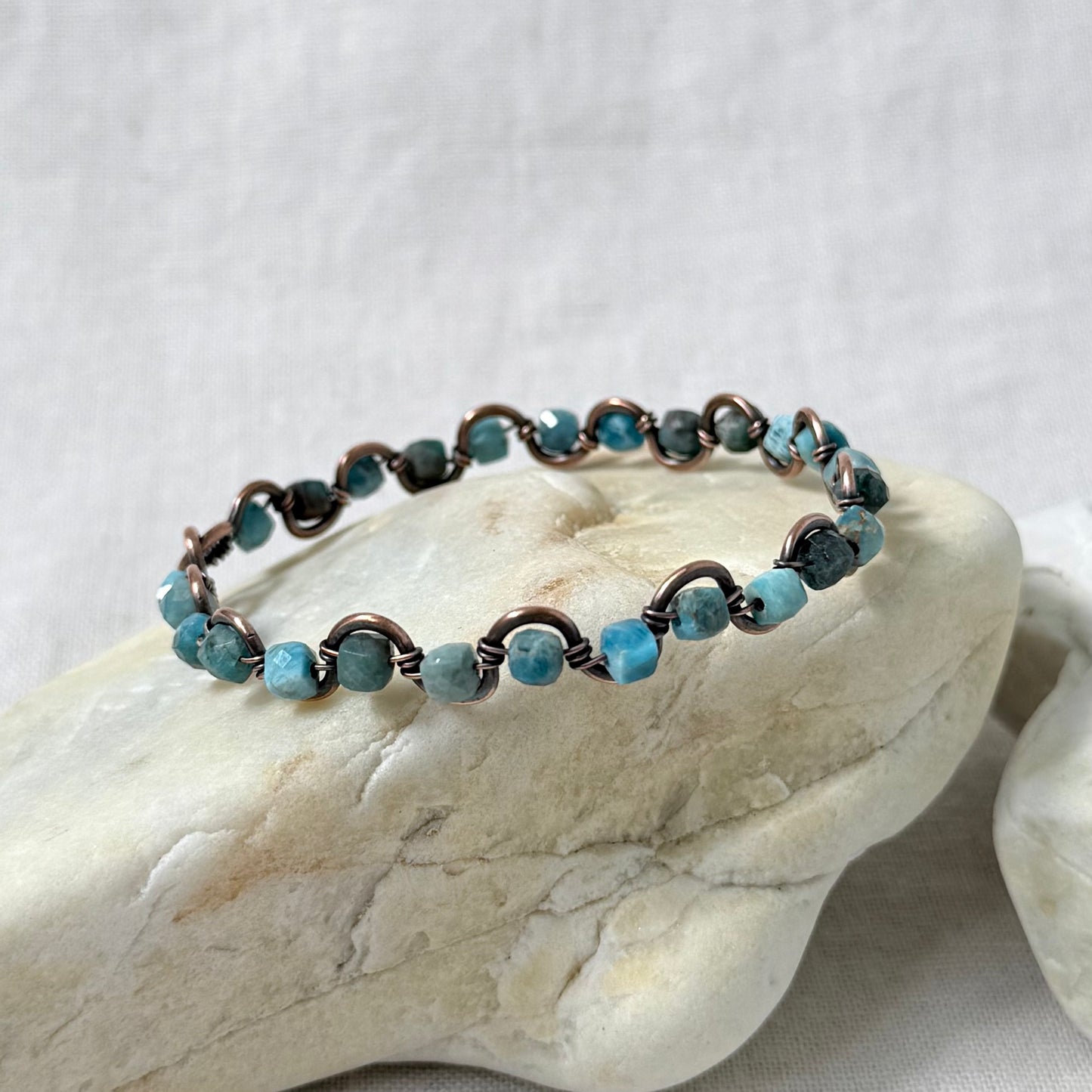 A delicate beaded copper bracelet with light blue Apatite beads, displayed on a light-coloured stone against a neutral fabric background.