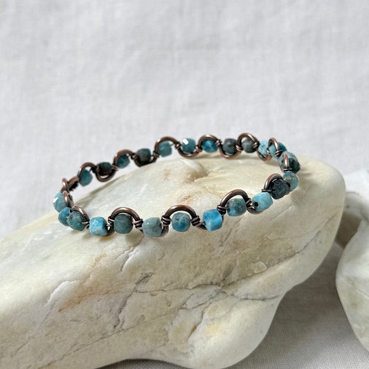 A delicate beaded copper bracelet with light blue Apatite beads, displayed on a light-coloured stone against a neutral fabric background.