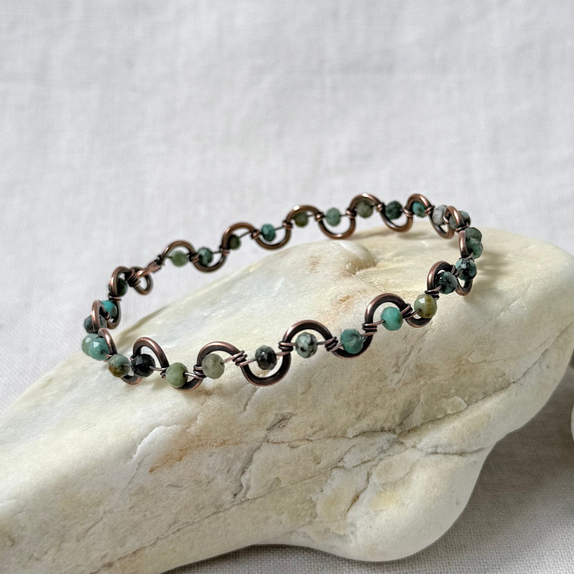 A delicate beaded copper bracelet with African turquoise beads, displayed on a light-coloured stone against a neutral fabric background.