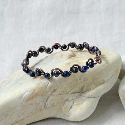 A delicate beaded copper bracelet with blue Lapis Lazuli beads, displayed on a light-coloured stone against a neutral fabric background.