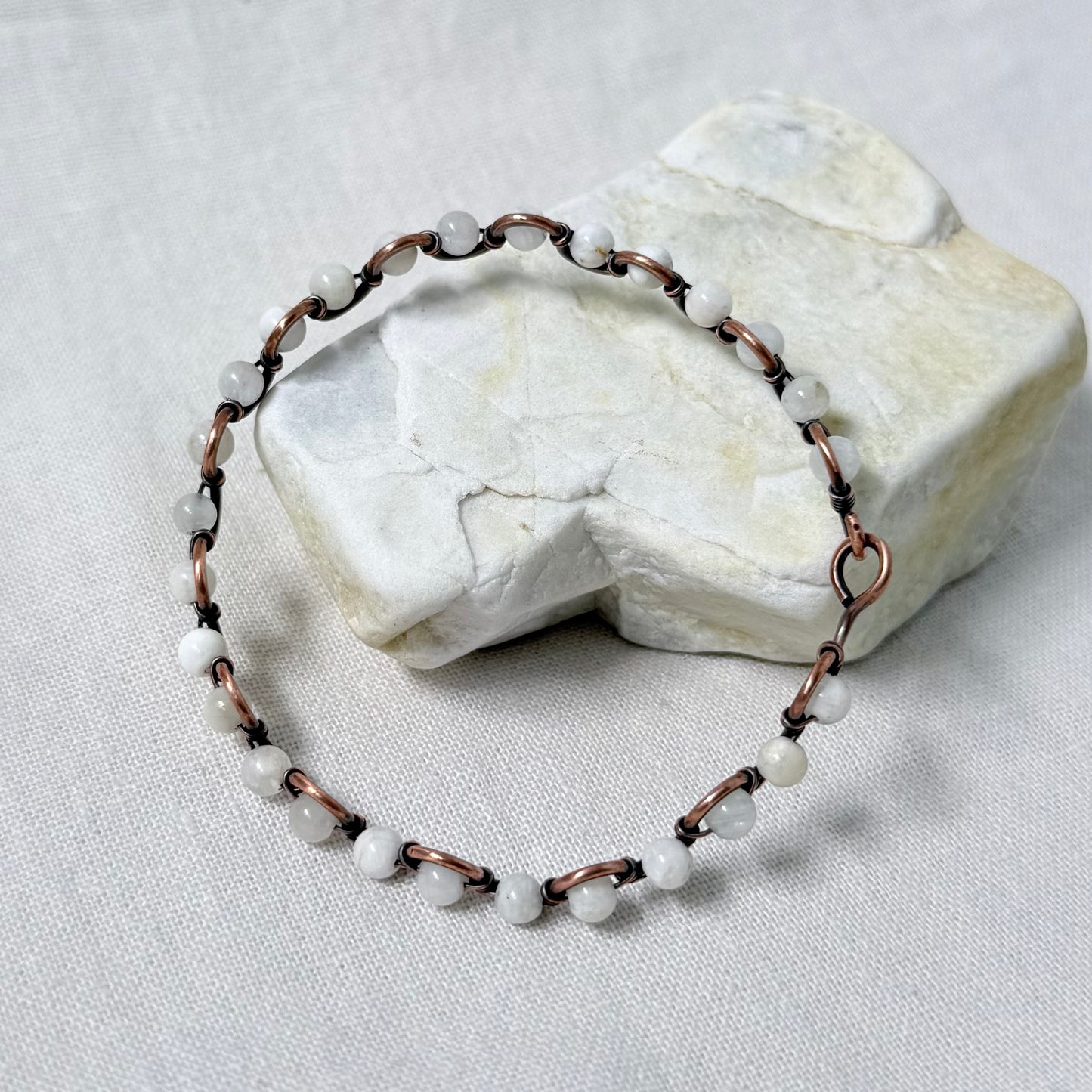 A handmade beaded bracelet with white round moonstone beads and copper wire, displayed on a textured white fabric surface with a white stone prop.