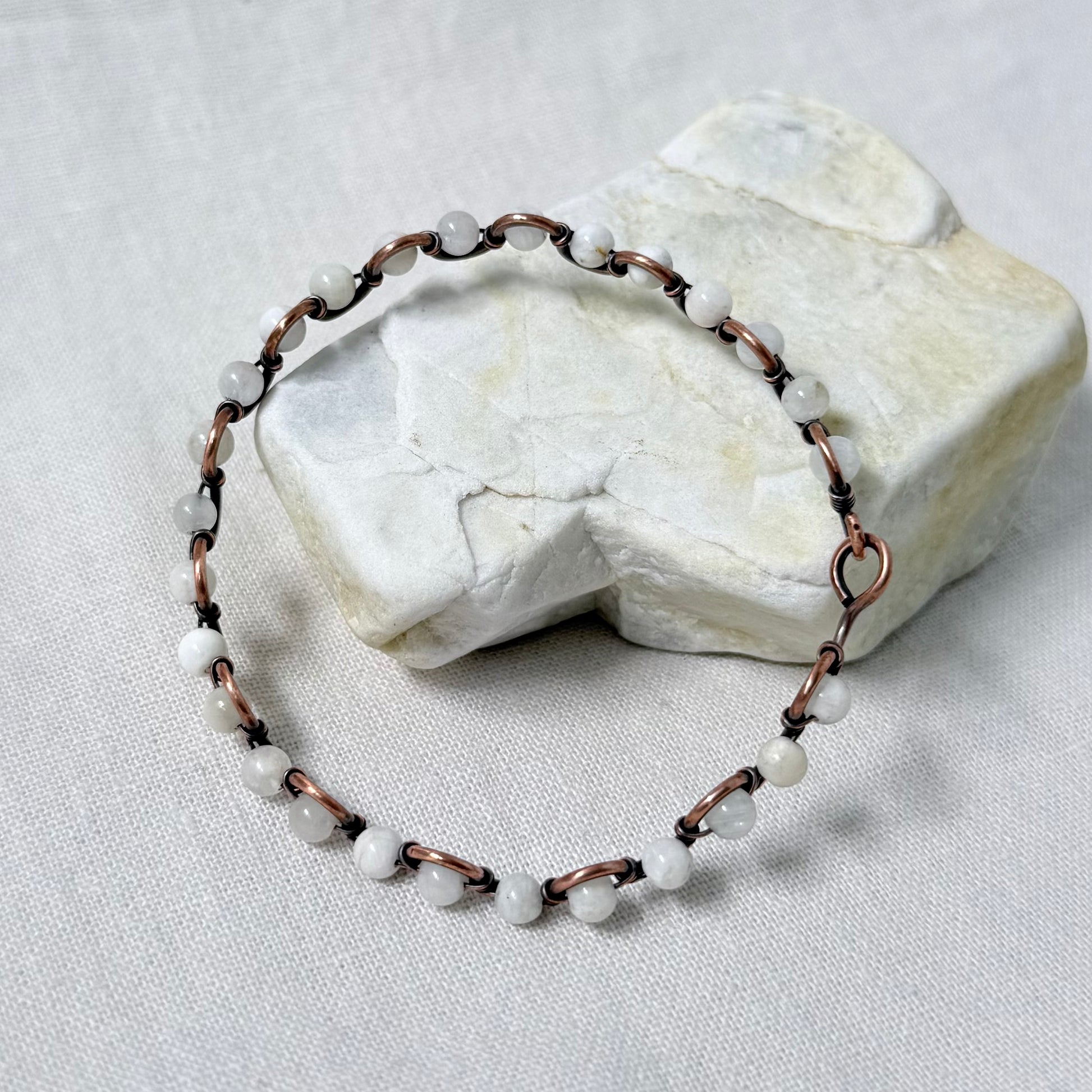 A handmade beaded bracelet with white round moonstone beads and copper wire, displayed on a textured white fabric surface with a white stone prop.