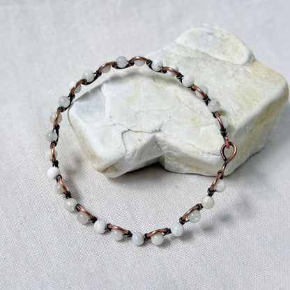 A handmade beaded bracelet with white round moonstone beads and copper wire, displayed on a textured white fabric surface with a white stone prop.