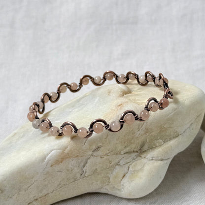 A delicate beaded copper bracelet with pale pink sunstone beads, displayed on a light-colored stone against a neutral fabric background.