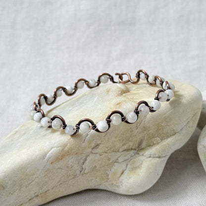A delicate beaded copper bracelet with white moonstone beads, displayed on a light-coloured stone against a neutral fabric background.