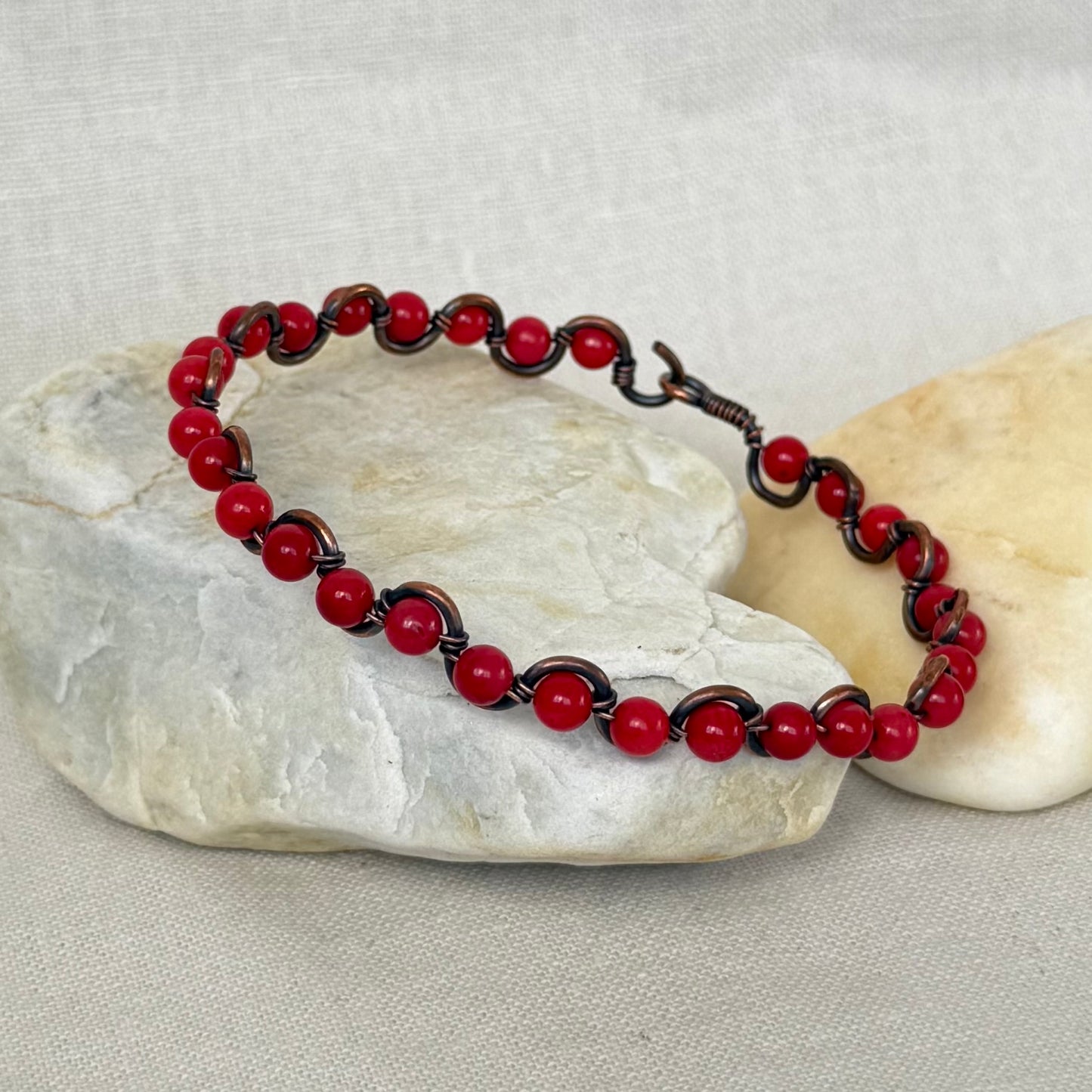 A beaded bracelet made of red coral round beads and a copper wire base, displayed on two smooth stones against a light fabric background.