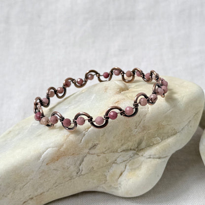 A delicate beaded copper bracelet with pale pink rhodonite beads, displayed on a light-coloured stone against a neutral fabric background.