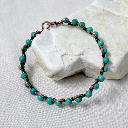 A delicate beaded copper bracelet with sinking turquoise beads, displayed on a light-coloured stone against a neutral fabric background.