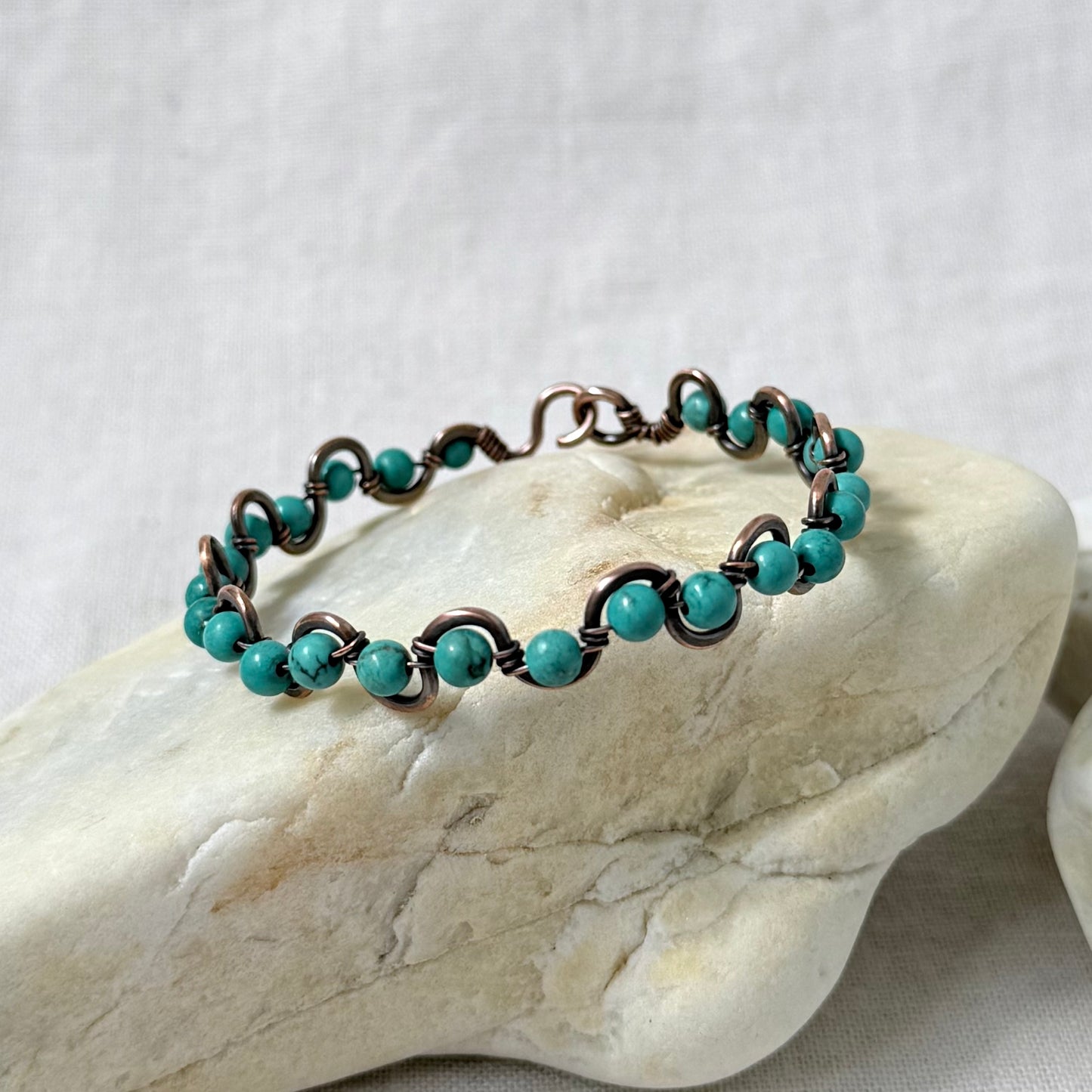 A delicate beaded copper bracelet with sinking turquoise beads, displayed on a light-coloured stone against a neutral fabric background.