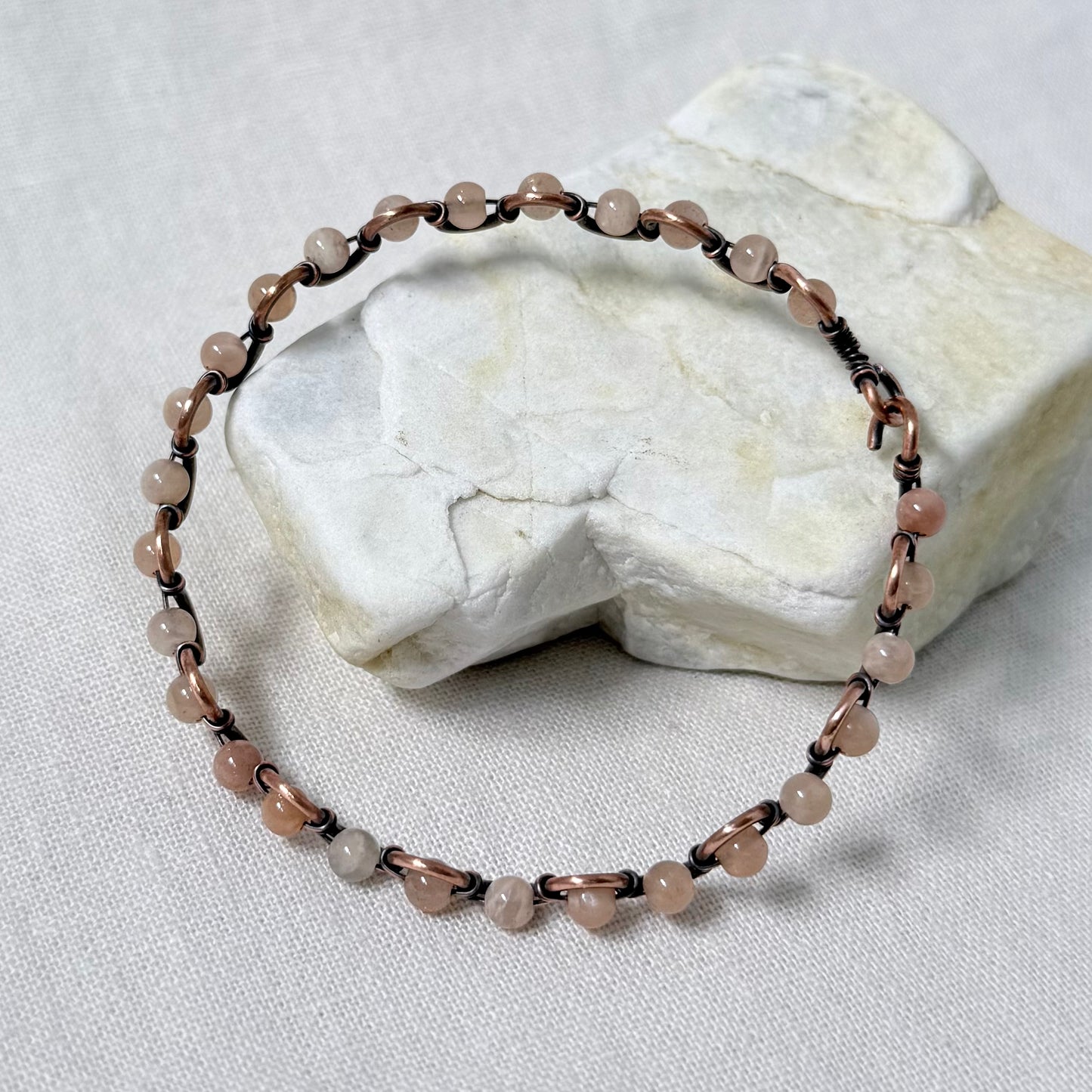 A beaded bracelet made of small sunstone beads and dark copper wire, displayed on a textured white stone against a white fabric background.