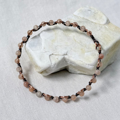 A beaded bracelet made of small sunstone beads and dark copper wire, displayed on a textured white stone against a white fabric background.