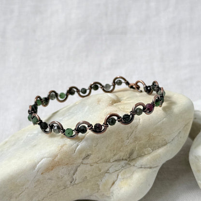 A delicate beaded copper bracelet with green Zoisite beads, displayed on a light-coloured stone against a neutral fabric background.