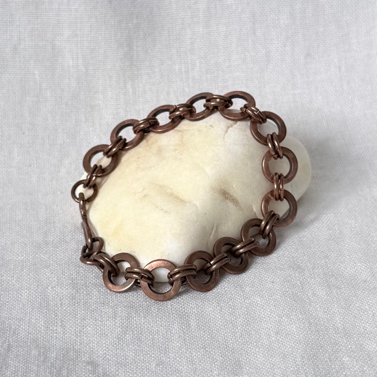 A copper chain bracelet with interlocking circular links, displayed on a smooth off-white stone against a light grey fabric background.