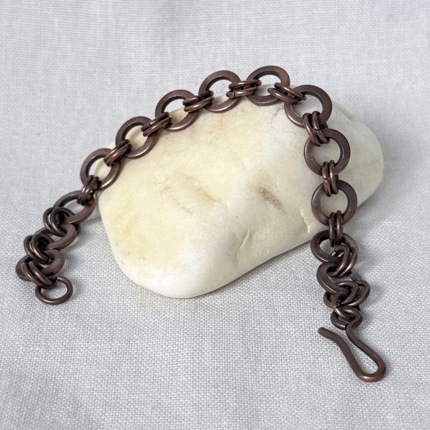 A copper chain link bracelet with a spring ring clasp, displayed on a smooth, light-coloured stone over a textured linen fabric background.