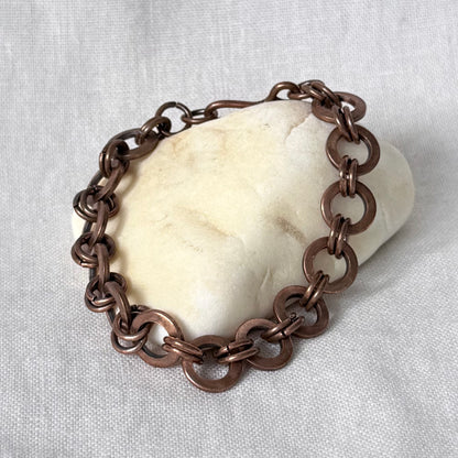 A copper chain link bracelet displayed on a smooth, off-white stone against a textured light grey fabric background.