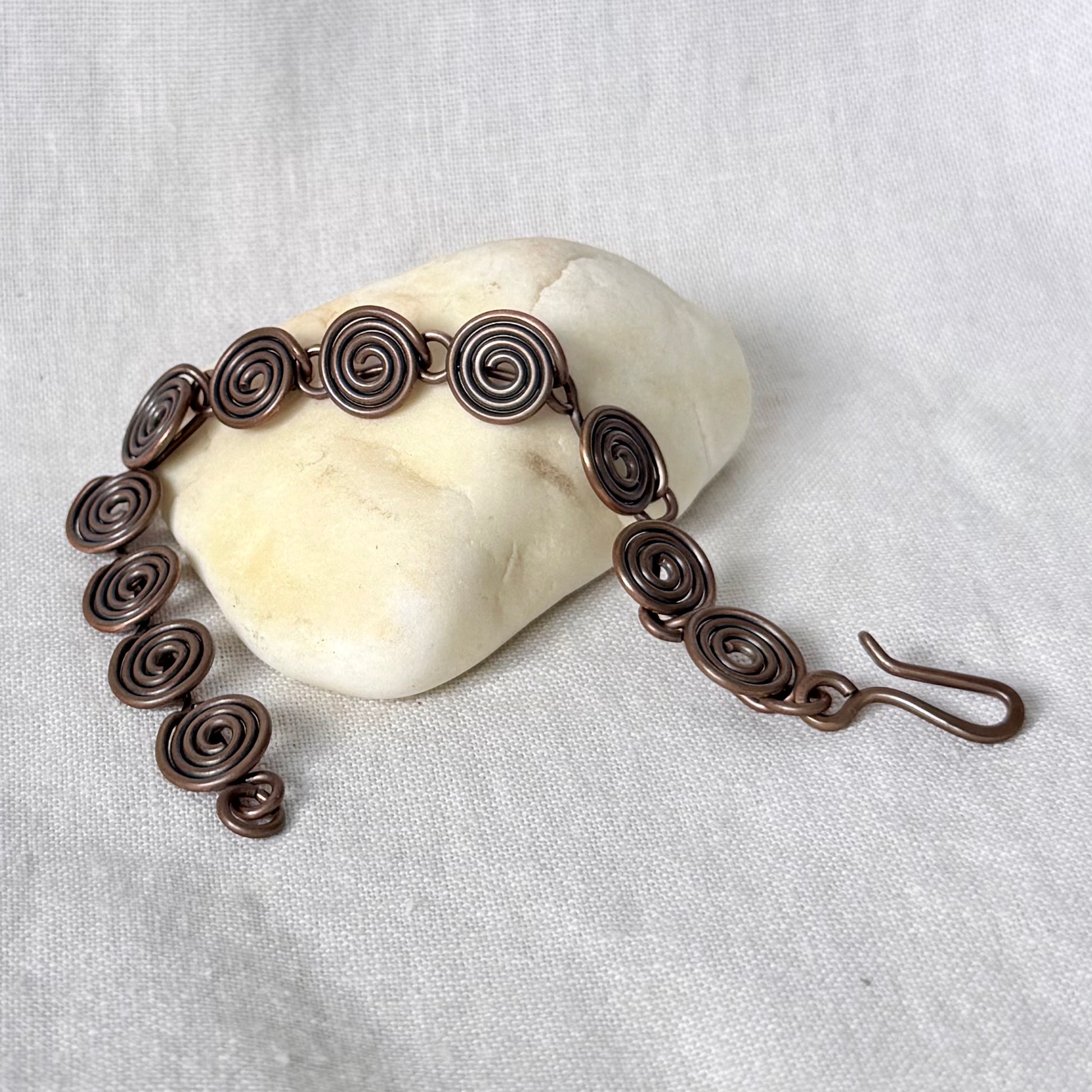 A handmade copper wire bracelet with spiral links, displayed on a smooth white stone against a textured off-white fabric background.