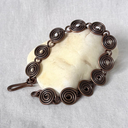 A handmade copper wire bracelet with spiral links, displayed on a cream-colored stone against a textured white linen background.