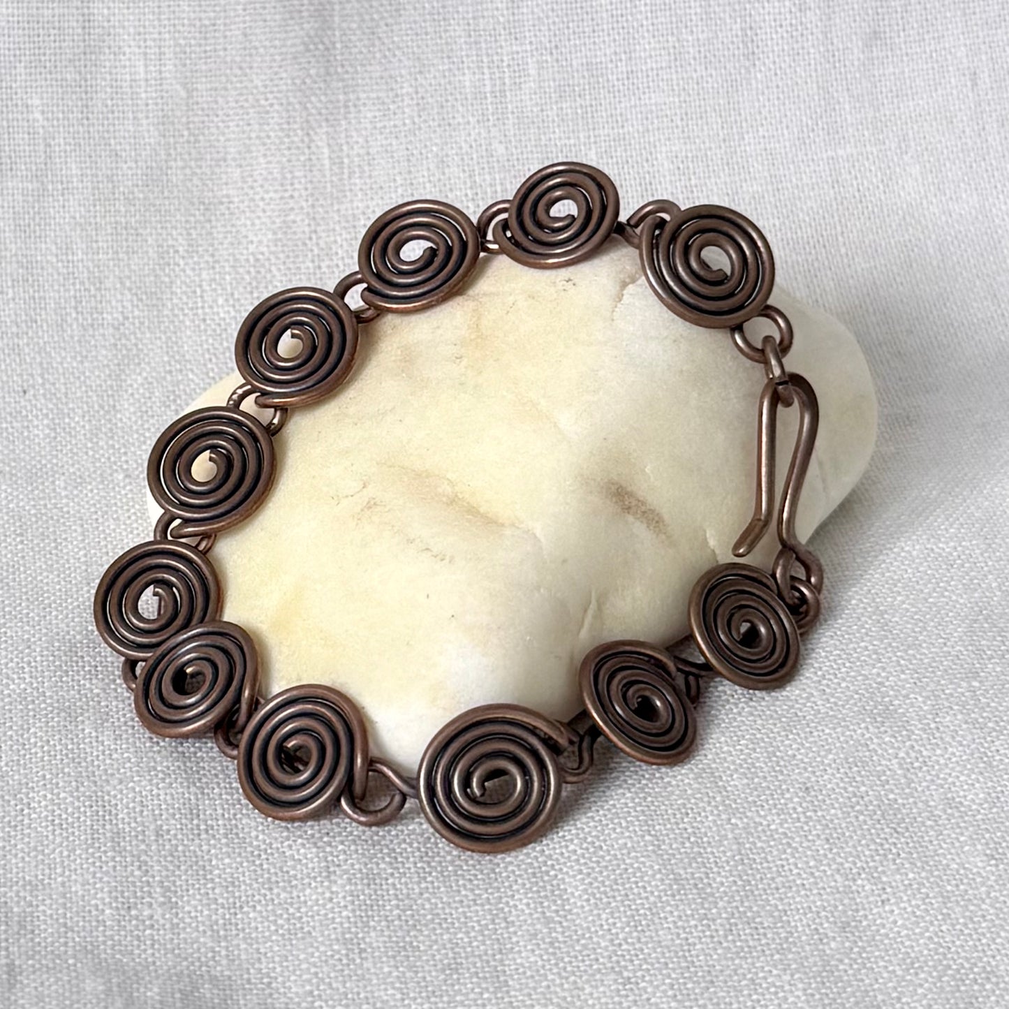 A handmade copper spiral link bracelet displayed on a smooth, off-white stone against a textured white fabric background.