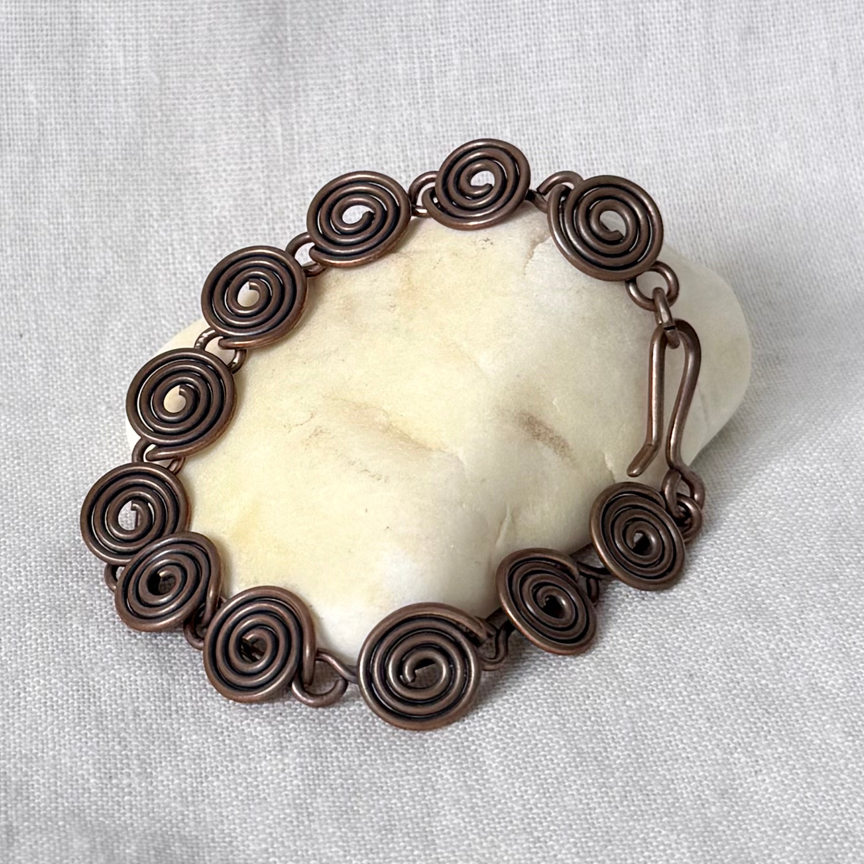 A handmade copper spiral link bracelet displayed on a smooth, off-white stone against a textured white fabric background.