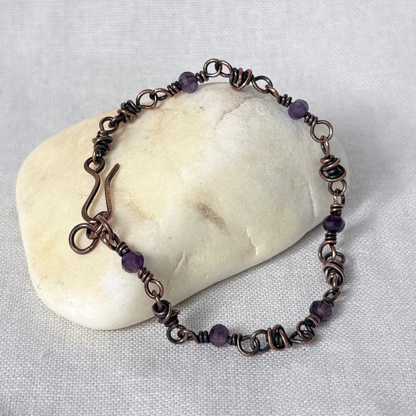 A handmade copper wire beaded bracelet with purple amethyst beads, displayed on a smooth cream-colored stone against a textured white fabric background.