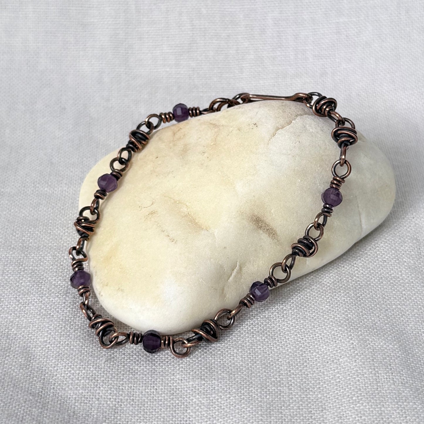 A handmade copper wire-wrapped bracelet with small purple amethyst beads, displayed on a smooth white stone over a light grey fabric background.