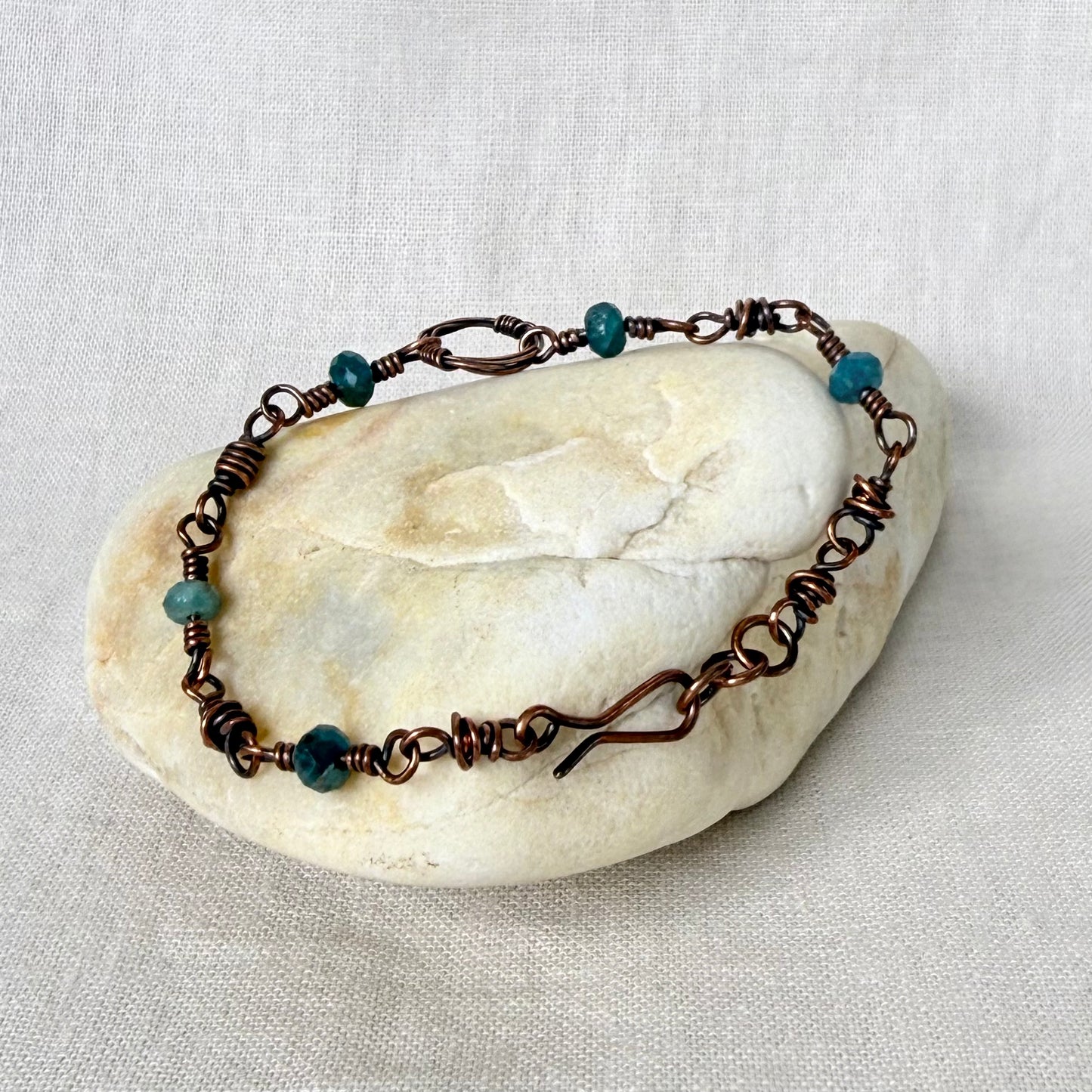 A handmade copper wire-wrapped bracelet with small teal beads, displayed on a smooth, light-colored stone against a neutral linen fabric background.