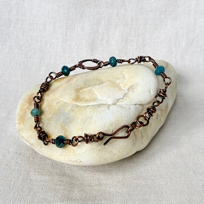 A handmade copper wire-wrapped bracelet with small teal beads, displayed on a smooth, light-colored stone against a neutral linen fabric background.